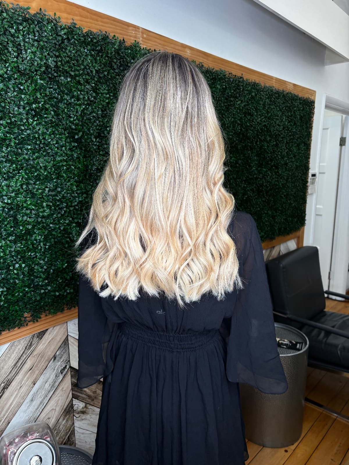 Waist-length blonde balayage with a deep shadow root melting into pale, sun-bleached ends, finished in loose beach waves against the salon's green boxwood wall — by Hali at McAllister Spa in Miami Beach