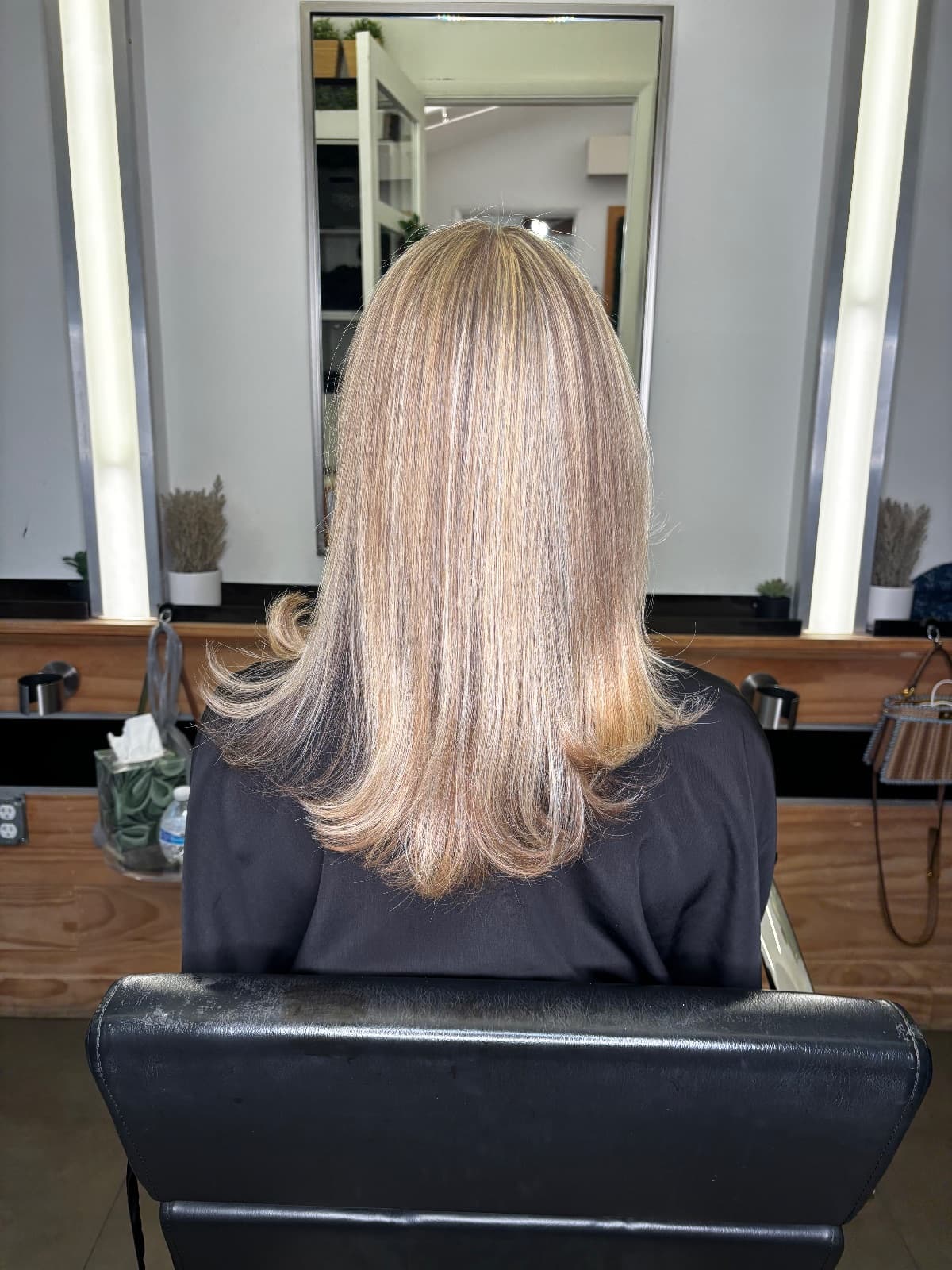 Shoulder-length beige blonde blowout with the ends flicked outward, photographed in the styling chair facing the lit mirror — highlights by Hali at McAllister Spa in Miami Beach