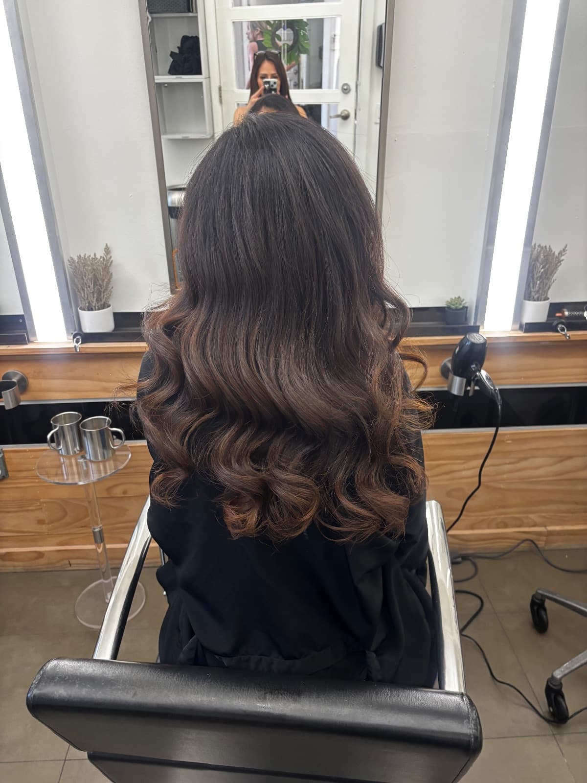 Dark brunette with warm chestnut balayage through the ends, set in wide bouncy curls, photographed in the chair with the stylist reflected behind — color and styling by Hali at McAllister Spa in Miami Beach